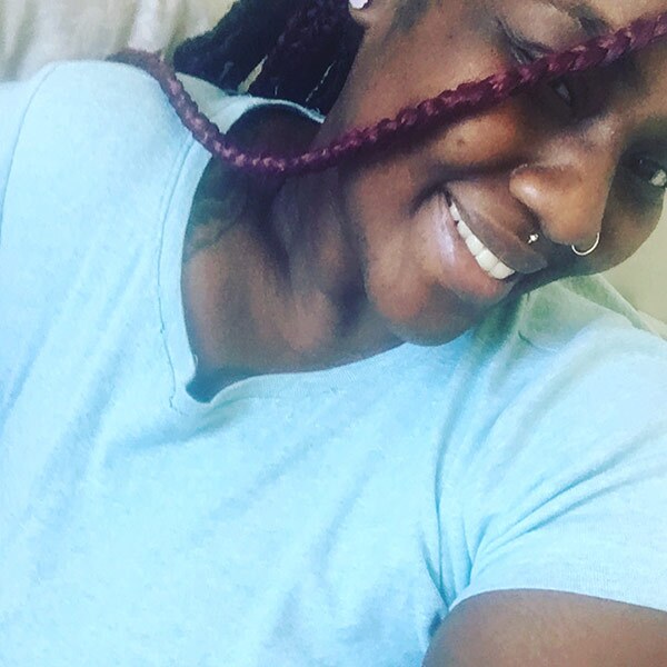 A young person wearing a light blue shirt smiles for a selfie as a braid falls over their face. 