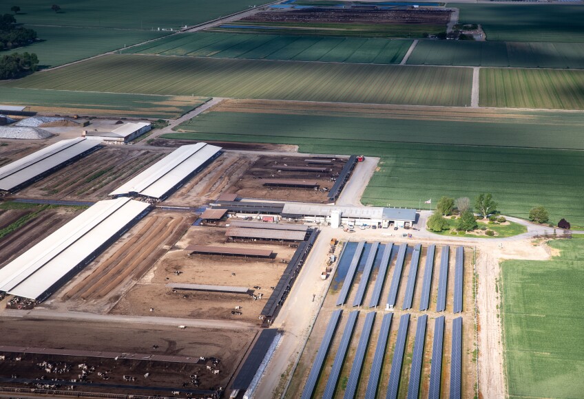 An aerial view shows the rectangular plots of green farmland and brown ranchland with cows visible in the bottom left corner and the landscape illuminated by the sun's rays with shadows cast by clouds.