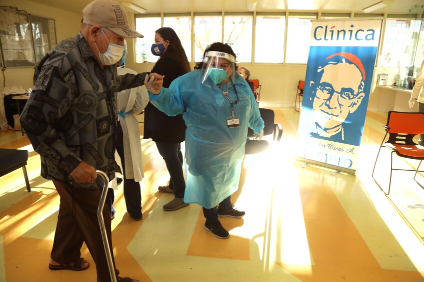 An elderly man wearing a baseball hat and a face mask and carrying a cane gets a helping hand from a health worker wearing PPE inside a medical clinic with sunlight streaming through a row of windows