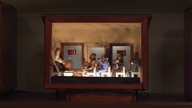 A shadowbox diorama with a wooden frame depicts a scene of clay figures sitting in chairs, with their attention pointed towards a miniature Malcom X figure at a podium. Malcolm X is teaching at the podium and the environment implies that they are in a mosque. The diorama is lit from below, casting shadows the clay figures on the upper part of the shadowbox.