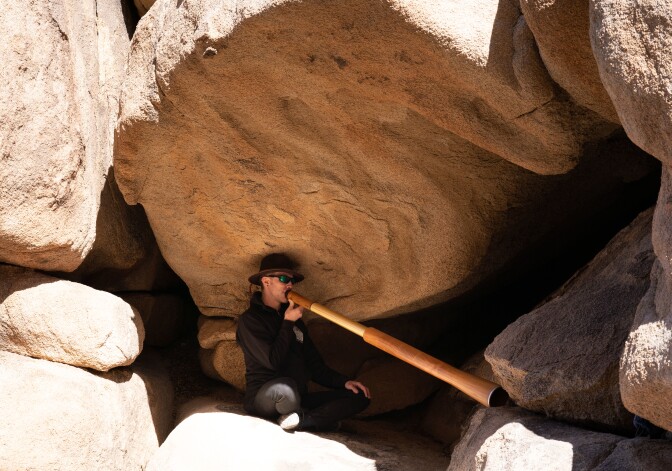 David Yates sits in a small crook in between large rock formations, as he plays the didjeridu — a long, wind instrument. The didjeridu is pressed up to his lips and the other end is propped up on a rock. Yates is sitting in the shade, provided by the rock formation above him.
