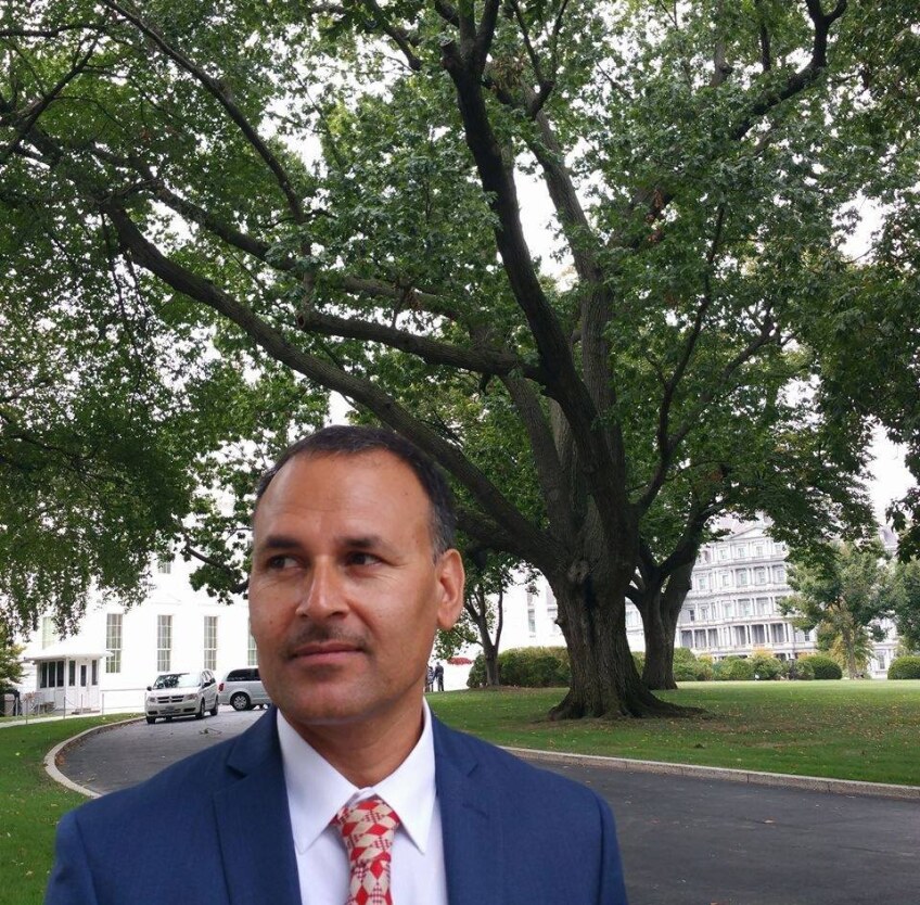 A man in a blue suit and red and gold tie stands in front of a tree.