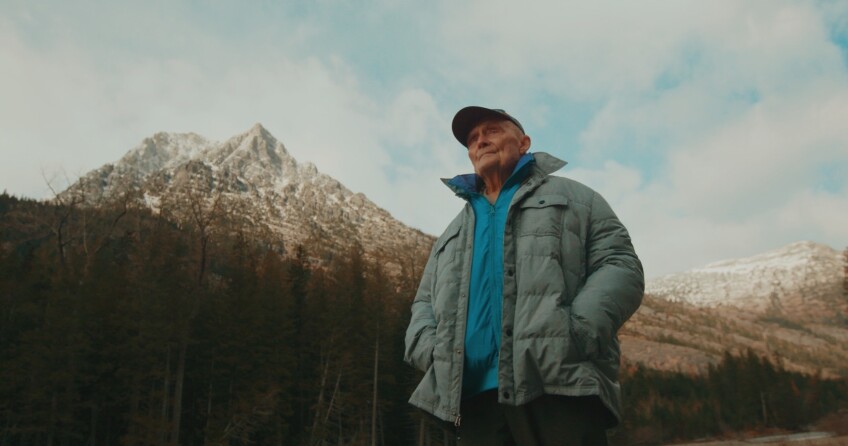 An elderly man stands wearing a powder blue puffer jacket and a teal windbreaker underneath. His hands are in his pockets and a baseball cap sits on top of his head. Behind him is a gorgeous landscape consisting of a towering, snowcapped mountain to his left and a forest of trees of various shades of greens, orange and yellow.