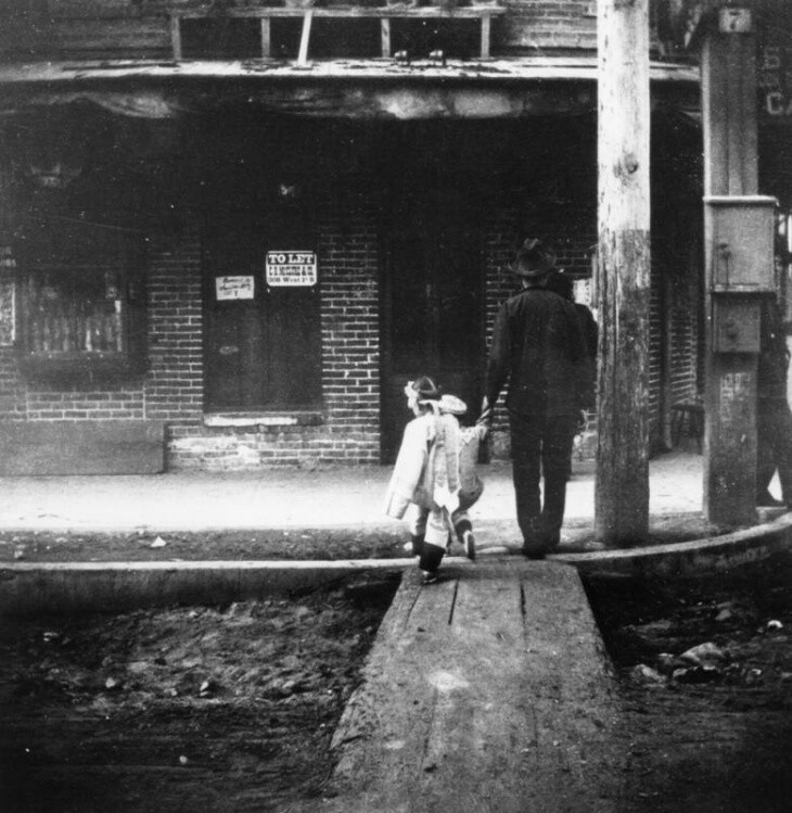 A man assists a child across the street on wooden planks in L.A.'s Old Chinatown, circa 1930s. | Los Angeles Public Library Collection