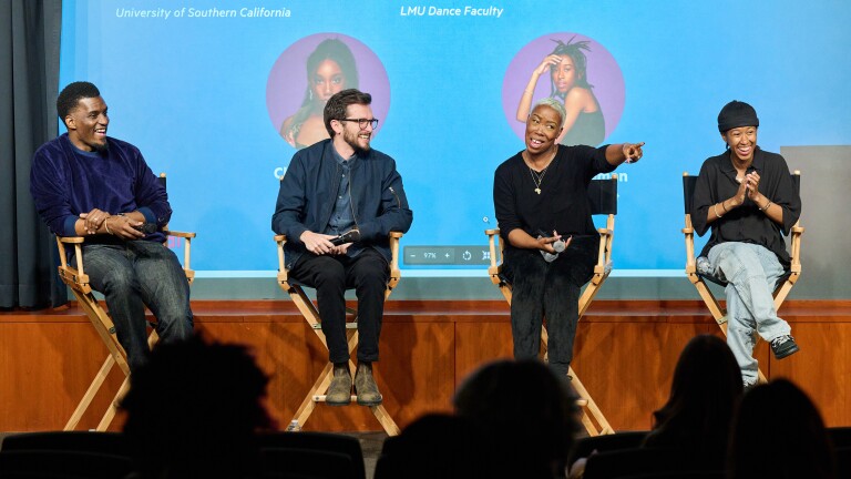 A panel of four people sit in director's chairs with amused expressions, and the third person from the left points to the crowd.