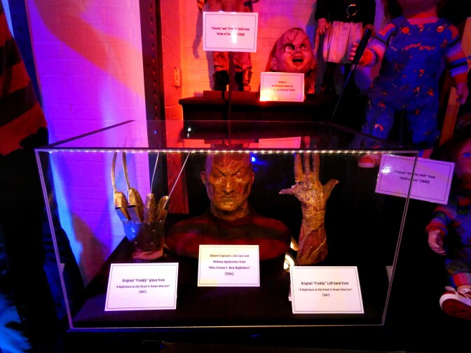 The original Freddy Krueger glove, hand and a life cast of Robert Englund on encased in a glass box. The glove is mechanical, with sharp blades coming out of each knuckle. The hand is mangled, resembling burned flesh. The life cast is a face mold depicting a menacing face.