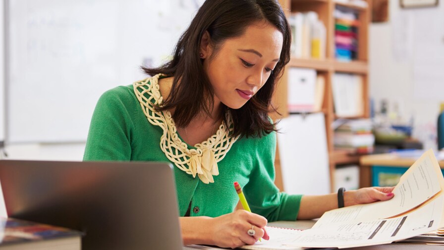 A woman in a green sweater looks down at some papers as she holds a pen. A laptop can be seen next to her.