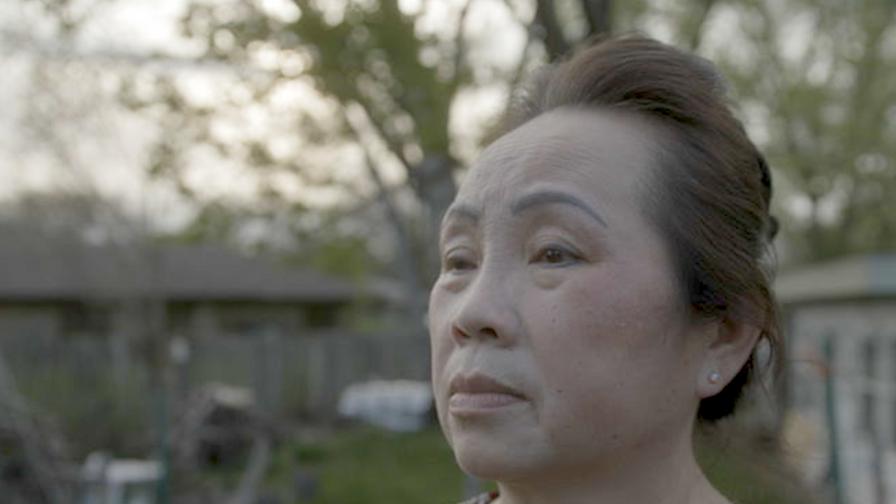 A photo of Youa Vang Lee, taken from the neck up. She has a solemn look on her face as she looks to at something out of frame. She is standing in a backyard, the background slightly blurred due to the depth of field.