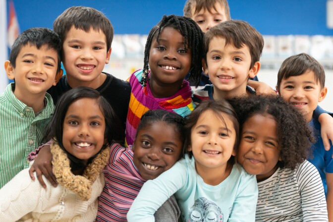 A group of diverse kids smile in this portrait. They are stacked on top of each other while cuddling in close and showing how happy they are.