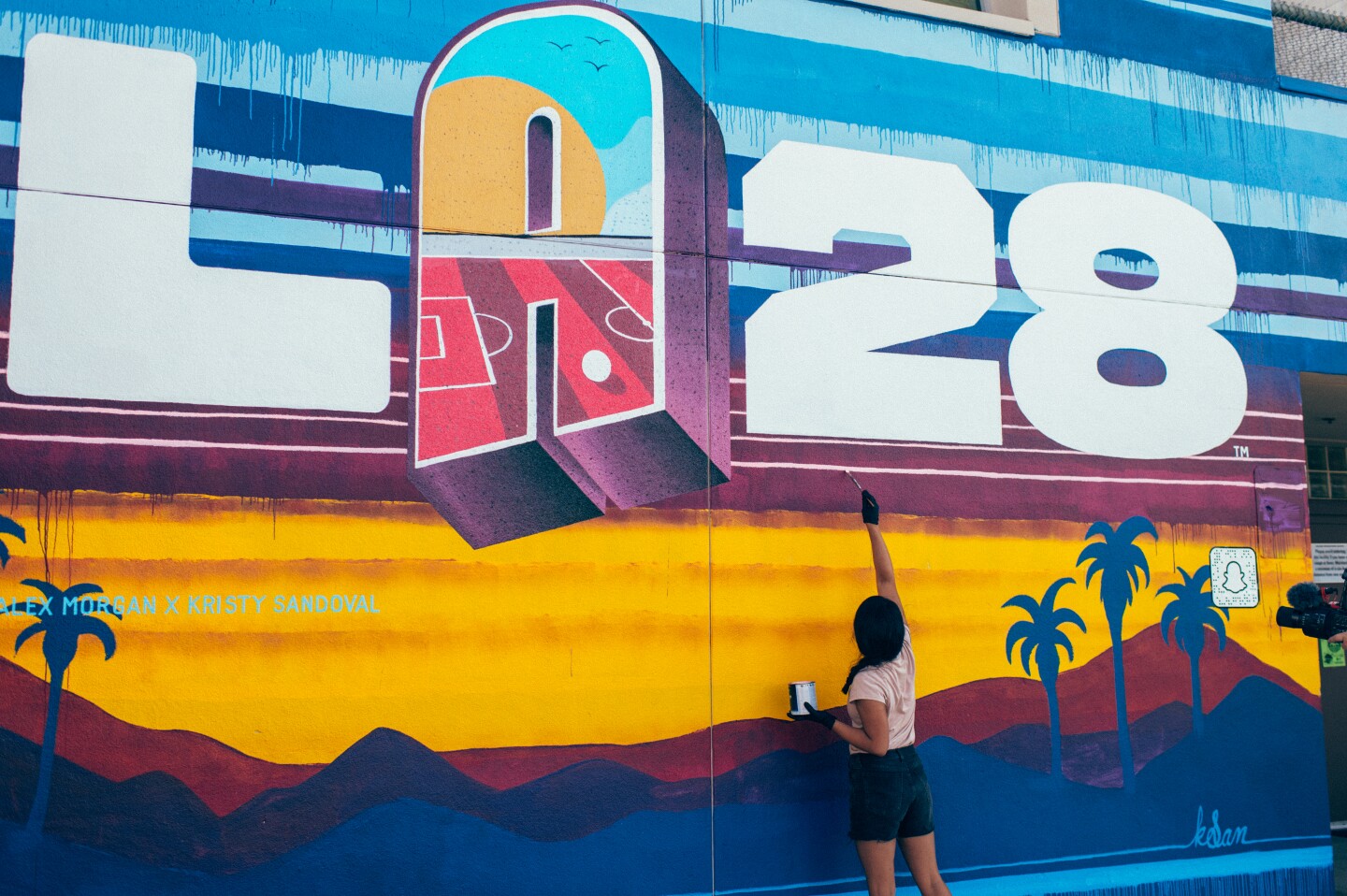 A young woman paints a a mural that says "LA28" with a sunset and palm tree silhouettes