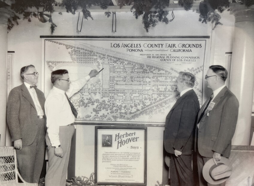 A black and white photo of four old, white men standing in front of future plans for the Pomona fair. The plans include an aerial drawing of what the fairgrounds would look like. At the top right corner of the plan, which is mounted on the wall, is "Los Angeles County Fair Grounds, Pomona, California." One of the men is pointing at a portion of the plan while the other three men look on.