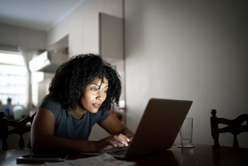 A Black woman with curly hair sits at a table in front of a laptop, with her face illuminated by the lit screen