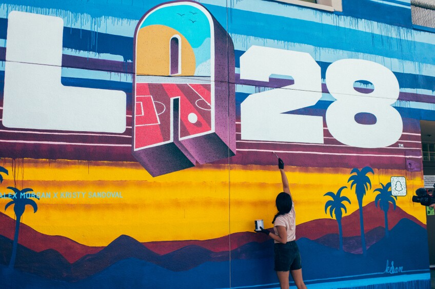 A young woman paints a a mural that says "LA28" with a sunset and palm tree silhouettes 