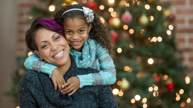 A Black woman and a small child hug each other and smile in front of an unfocused Christmas tree.