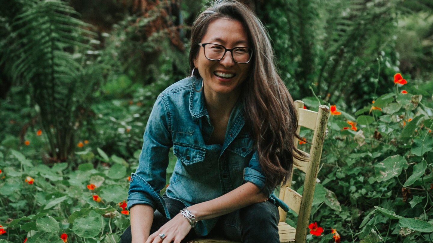 A woman with long hair and a denim jacket sits on a chair in a forest.