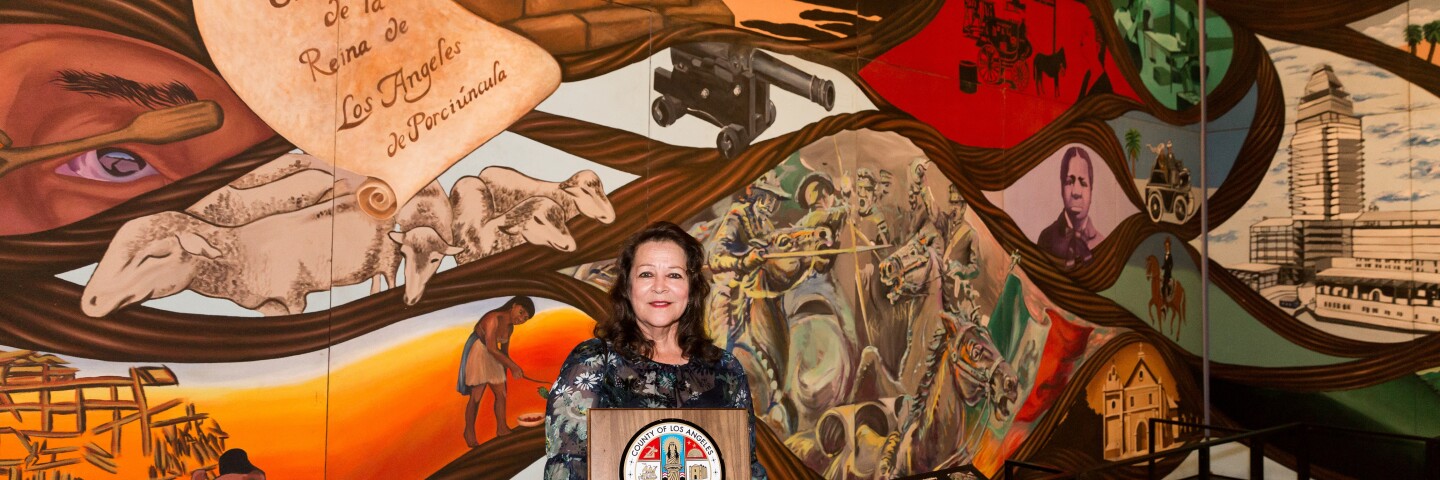 Artist Barbara Carrasco standing in front of colorful floor-to-wall length mural "L.A. History: A Mexican Perspective" holding a commendation plaque from Los Angeles County presented by County Supervisor Hilda Solis at the opening of the temporary exhibition "Sin Censura."