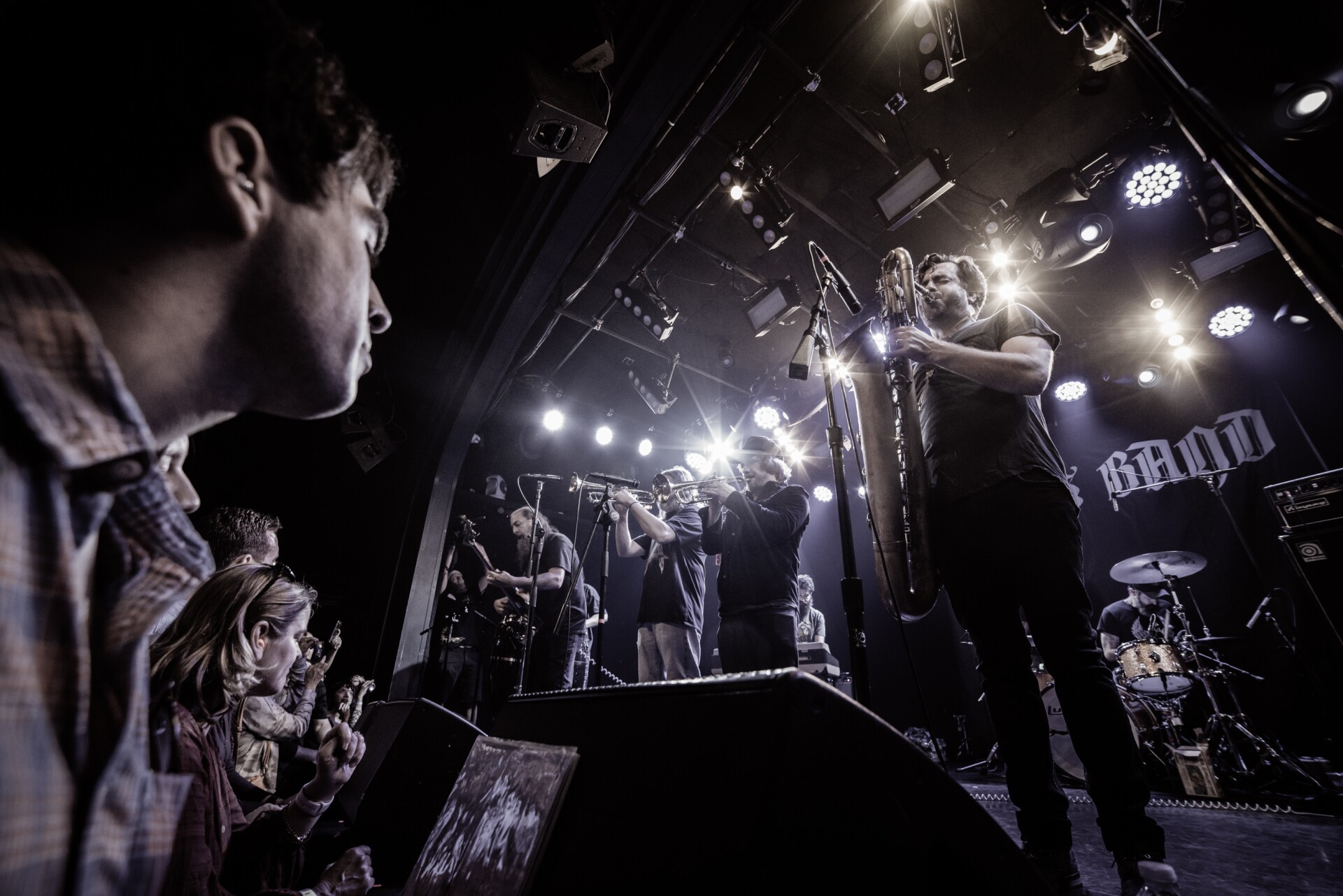 A band performs on a stage. From left to right there is a guitarist, two trumpet players and a saxophonist. The audience listens to the left of the image. Behind the band are bright white stage lights.