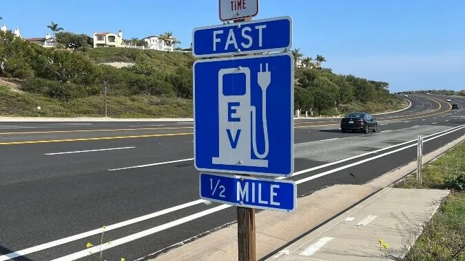 A car passes a blue and white EV charging station directional sign on a curved highway.