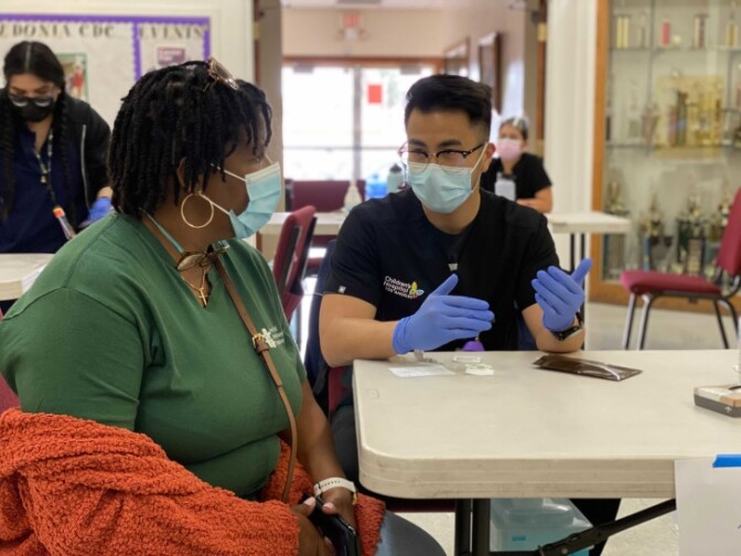 Edwina Shivers is wearing a green t-shirt and a bright orange sweater as she sits at a plastic foldable table. She's listening to David Nguyen in a black t-shirt and glasses as he explains something to her. He's also wearing medical gloves.