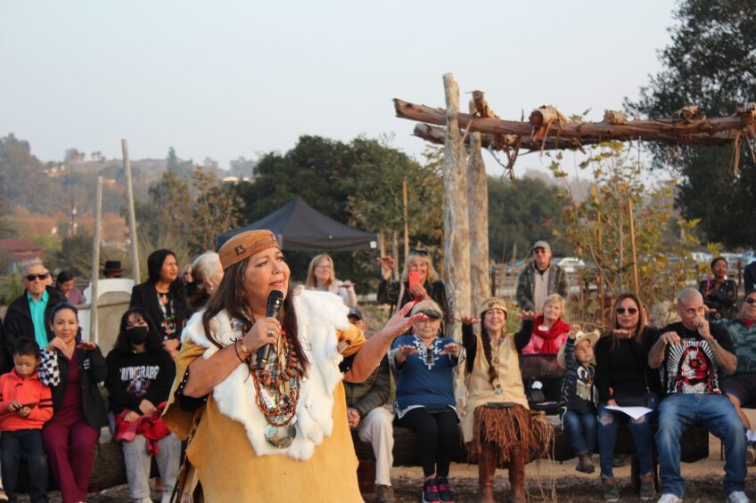 A woman in traditional Juaneno Acjachemen regalia stands in front of a crowd of people seated and gathered around her. She is holding a microphone and is speaking to a crowd, out of frame. The people seated behind her have their arms stretched out towards her and look happy.