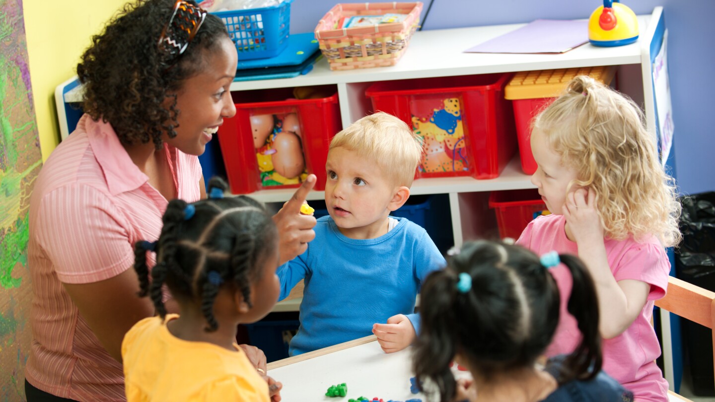 Small children at a preschool center. A teacher holds up one finger as she counts.