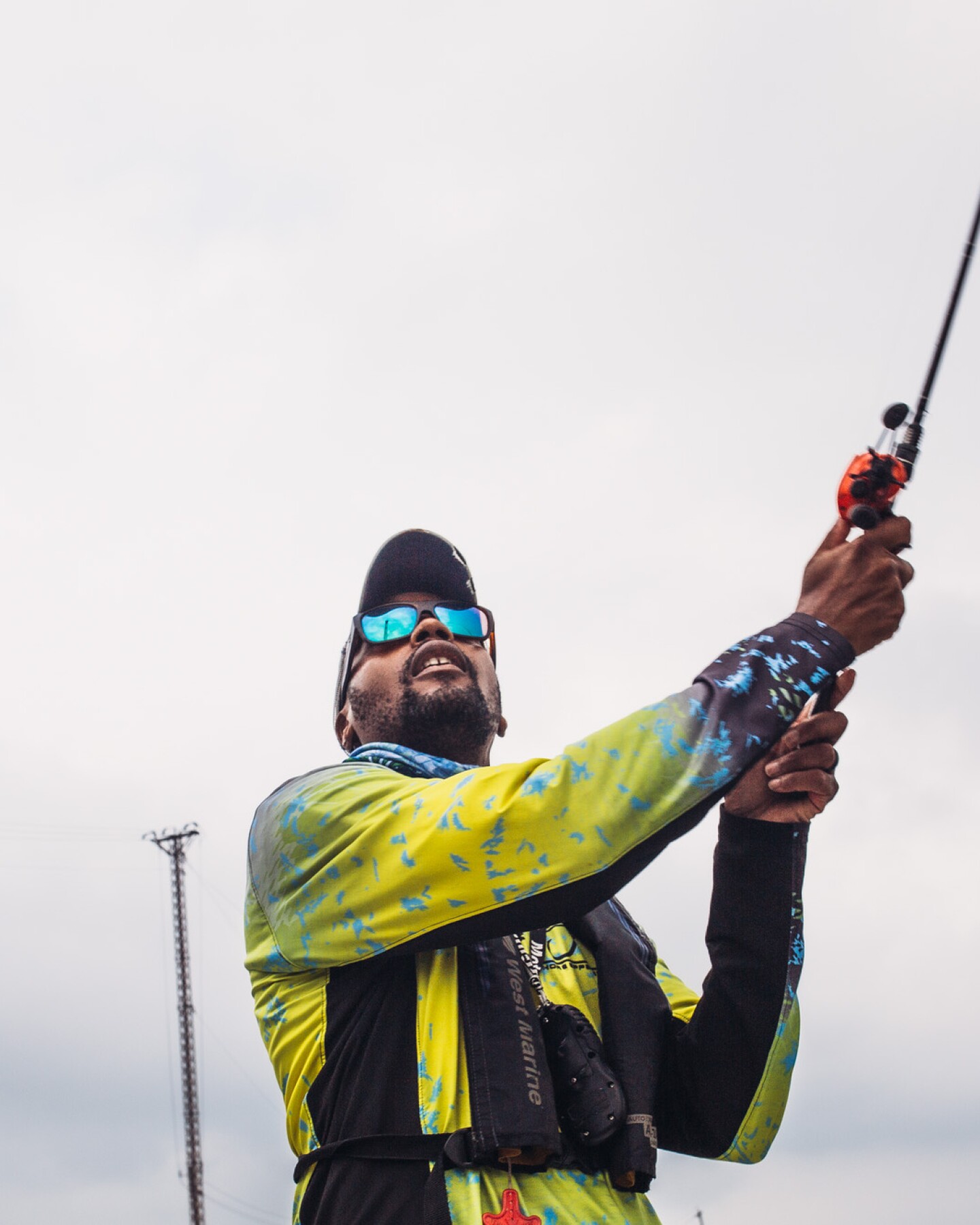 A man casts a fishing line. Behind him is a gloomy, gray-blue sky. The man is wearing sunglasses with teal blue lenses and a bright yellow jacket.