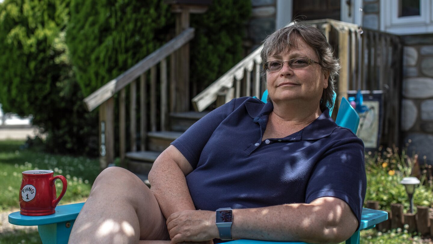 Susan sits on a bright blue wooden lawn chair. She's sitting leaned back, with her legs folded and her arms crossed. Patches of sunlight light different parts of her body as she smiles with a gaze slightly off from the camera.