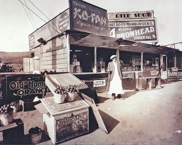 A sepia-tone historic photo of a man holding a cane standing in front of a food stand, surrounded by various crates, boxes and advertising signs promoting cigarettes, candies, barbeque and more.