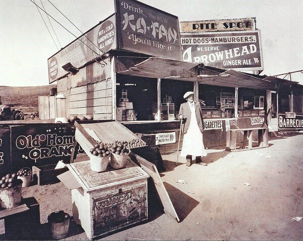 A sepia-tone historic photo of a man holding a cane standing in front of a food stand, surrounded by various crates, boxes and advertising signs promoting cigarettes, candies, barbeque and more.