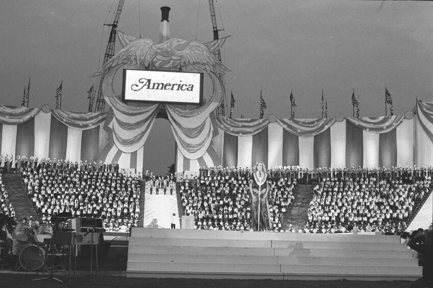 Singer on stage at LA Coliseum, "America" on electronic sign, patriotic decorations and full audience