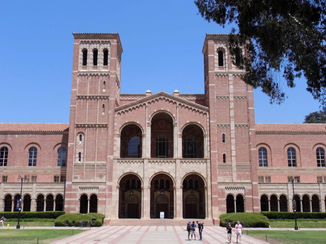 A red-brick building stands in an open quad with green grass and a paved walkway leading up to the steps of the building.
