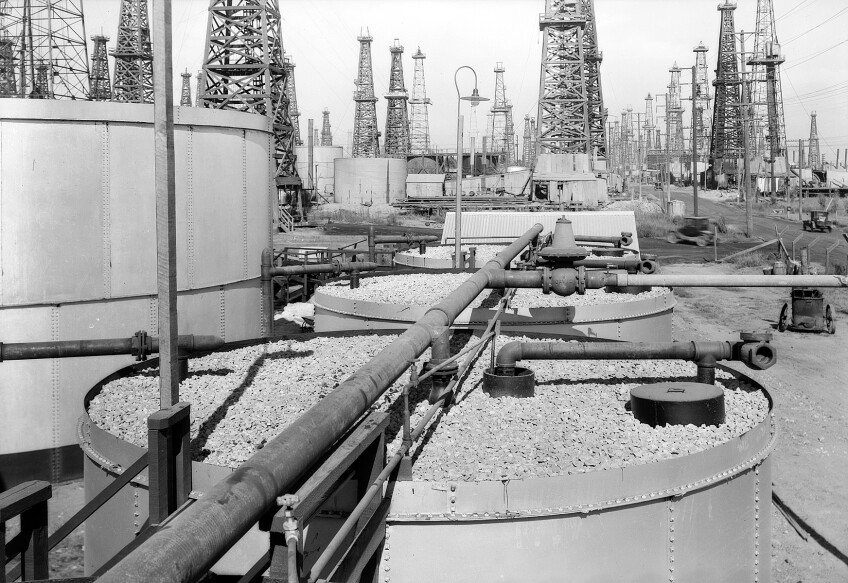 A black and white photo of oil tanks. Oil wells are lined up, stretching for miles beyond.