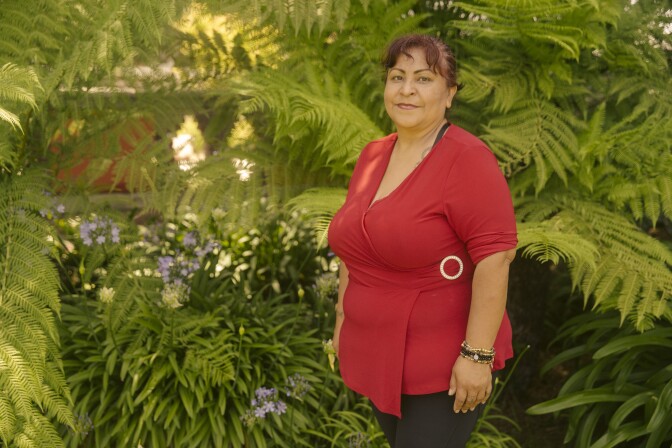 A woman in a red top stands and smiles in front of large plants
