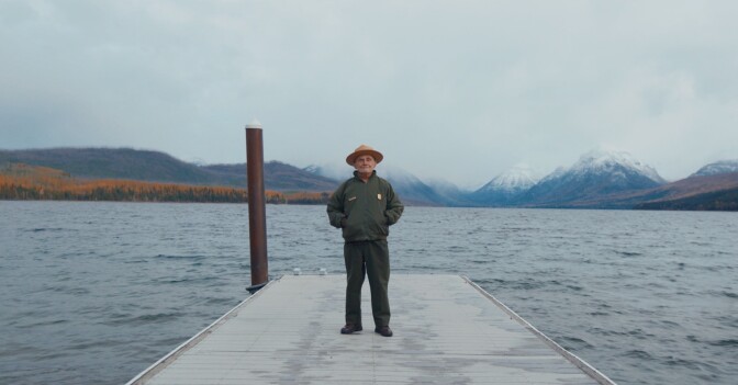 An elderly man wearing a broad-brimmed hat stands on a dock by a lake. Behind him are snow-capped mountains and a forest of orange-colored trees.
