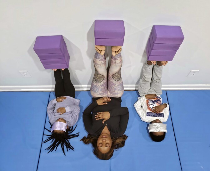 A black mother and her children practice a yoga pose with their legs resting on a wall as they close their eyes.