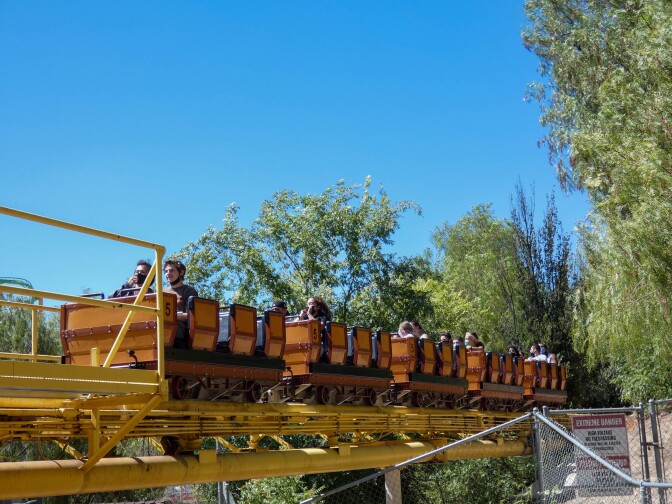 A row of rollercoaster cars meant to resemble Old West mine cars sit on a yellow track. Passengers are sat sporadically throughout the mine cars. The mine cars are mostly orange with red lining and details. Behind are tall, wispy green trees