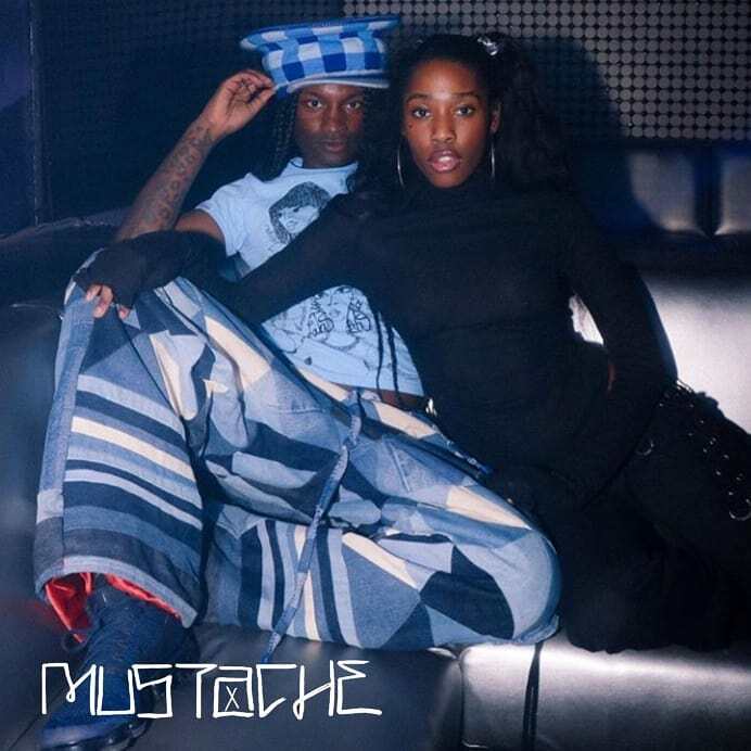 Two people, one in blue patterned wear and another in all black, are seated together.