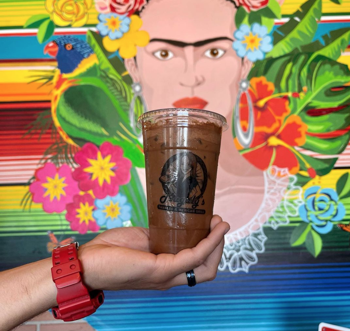 A tall plastic cup of iced coffee is held up. The iced coffee is a dark brown color and is held up against a bright and colorful mural of Frida Kahlo behind it.