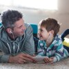 A man and a little boy watch something on a tablet together as they lie down on a carpet next to each other