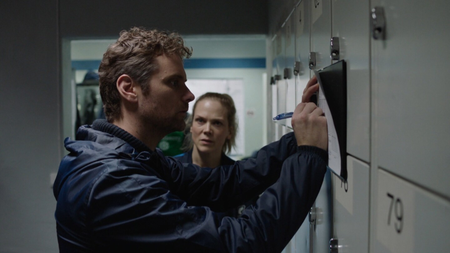 Helena faces Espen as he writes on a piece of paper against a locker door. | "Home Ground: Reserve Team"