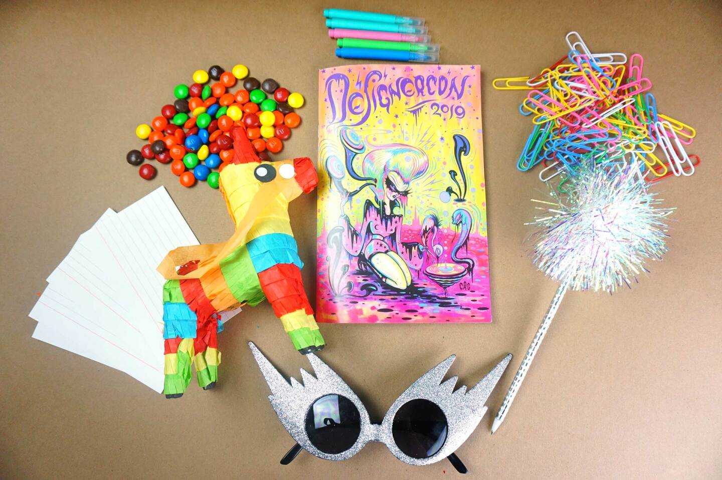Index cards, spiky sunglasses, a book, a small piñata, markers and colorful paper clips are laid out on a table.