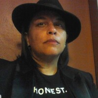 A headshot of SenYon Kelly. SenYon is wearing a black short-brimmed hat that is tilted slightly to one side. SenYon is looking direct to camera and is wearing a black shirt with a black collared shirt over. SenYon is also wearing hoop earrings and is standing in front of a brown wall background.