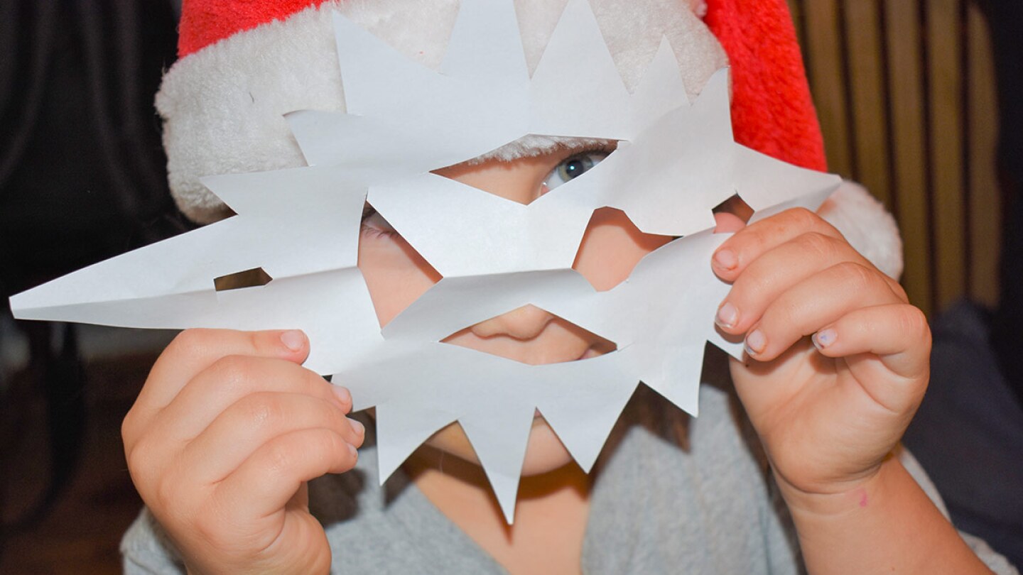 A small child wearing a red Santa hat holds a paper snowflake in front of the face like a mask.