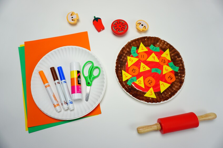 a pizza craft made of a paper plate made to look like a pizza using markers and construction paper shapes, which are lying next to the craft.