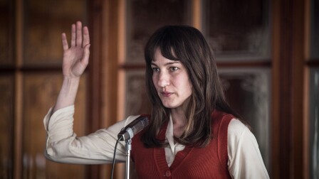 Marie Leuenberger as Nora raises her hand behind a mic stand.
