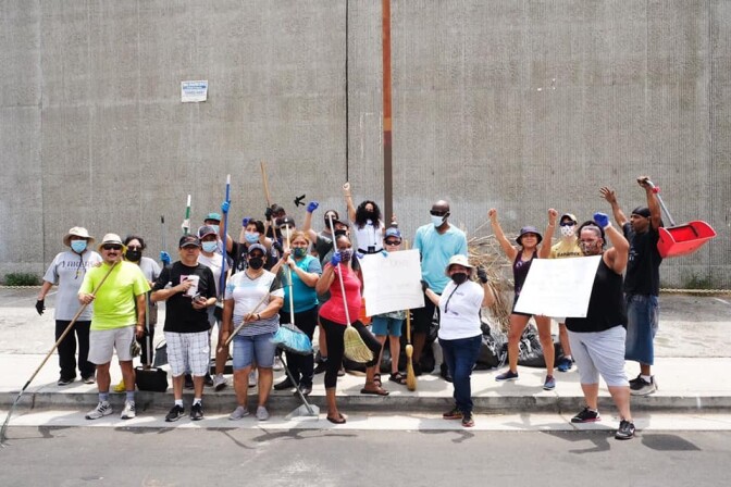 A group of people with raised fists, holding neighborhood cleanup supplies and signs to protest the developers demolishing their neighborhood.