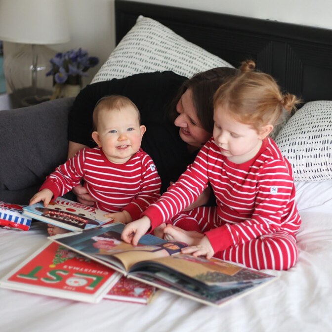A woman cuddles two small children in matching pijamas as they read together on a bed.