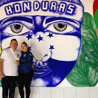 Lilian Meija and José Aguilar stand in front of a mural. The mural features a face painted in blue with strong, dark eyebrows, full lips and determined eyes. A band goes across the face's head with the words "Honduras" over it. To the left of the blue face is a multicolored guacamaya, or macaw. To the right of the blue face is half of a woman's face with large eyes, long eyelashes, full red lips and strong cheekbones. 