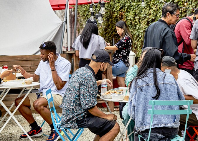 Attendees gather around outdoor tables to eat their purchases at the FilLed Market. Nearby is a tall wall covered in green vining plants.