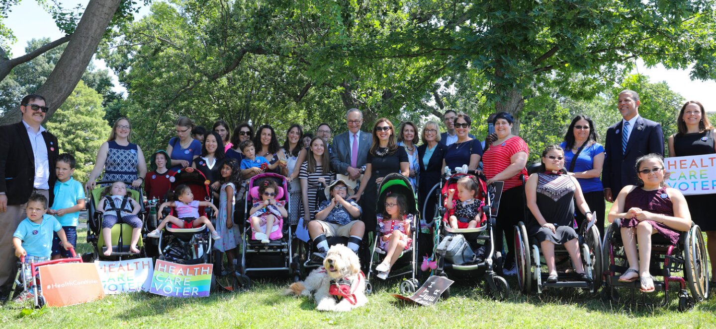 Disabled children and their parents with signs advocating for health care photographed in a green area